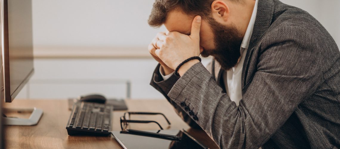 A businessman presses his hands over his eyes at his desk.